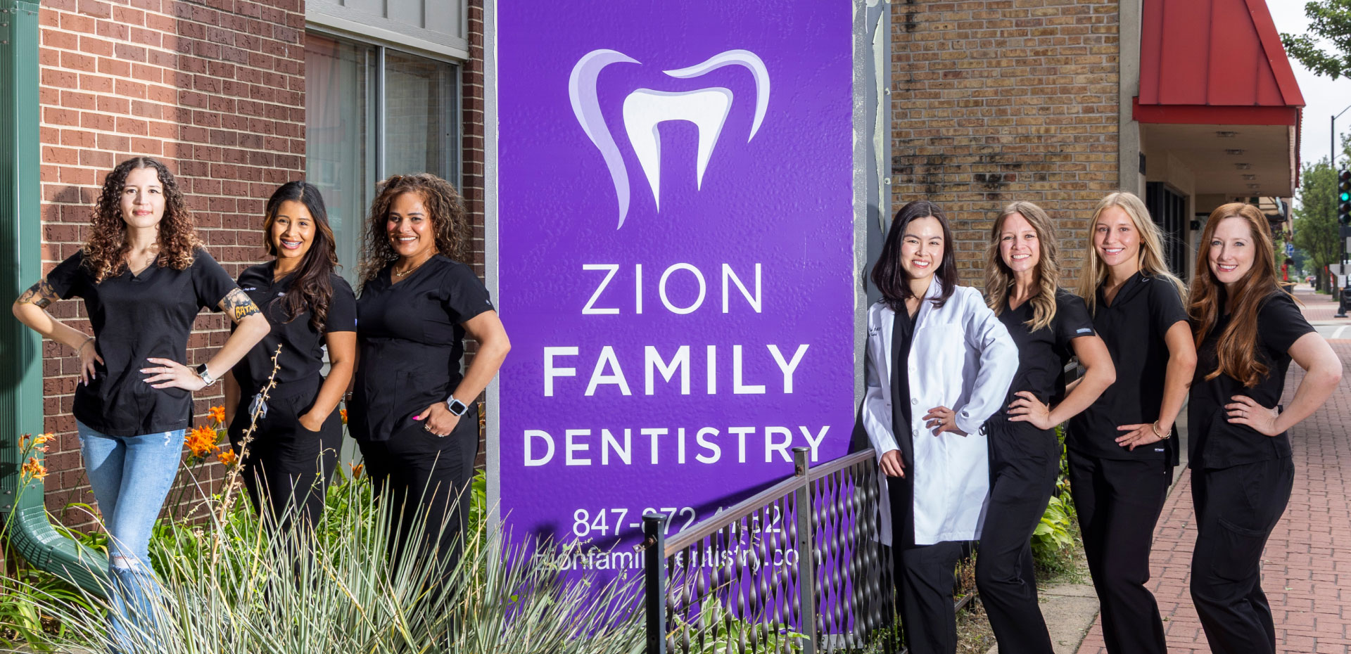 El equipo de Zion Family Dentistry posando junto al letrero de la oficina frente al edificio de ladrillo